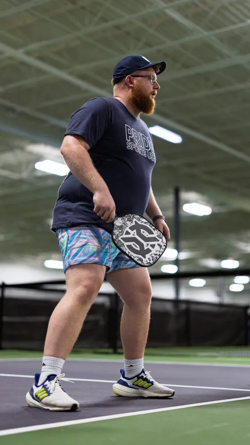 A pickleball player stands ready on an indoor court, holding a patterned paddle and wearing a "Pride Sports USA" shirt, colorful shorts, a cap, glasses, and athletic shoes. The background shows a net and bright overhead lights.