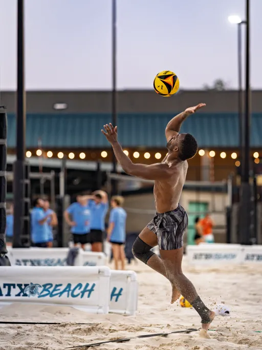 A volleyball player leaps off the sand to serve the ball, wearing striped shorts and a knee brace, on an outdoor sand volleyball court with other players and spectators in the background.