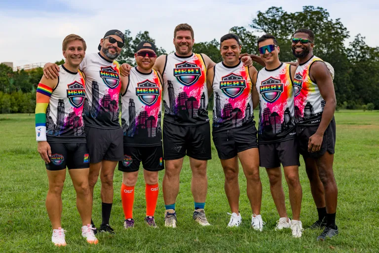 Seven ATX Heat (Orange) players pose arm-in-arm on a grassy flag football field during practice. They wear matching white jerseys with a pink‑orange HEAT shield and city skyline, black shorts, and athletic shoes; some have sunglasses, hats, or colorful armbands, with trees and open park space behind them.