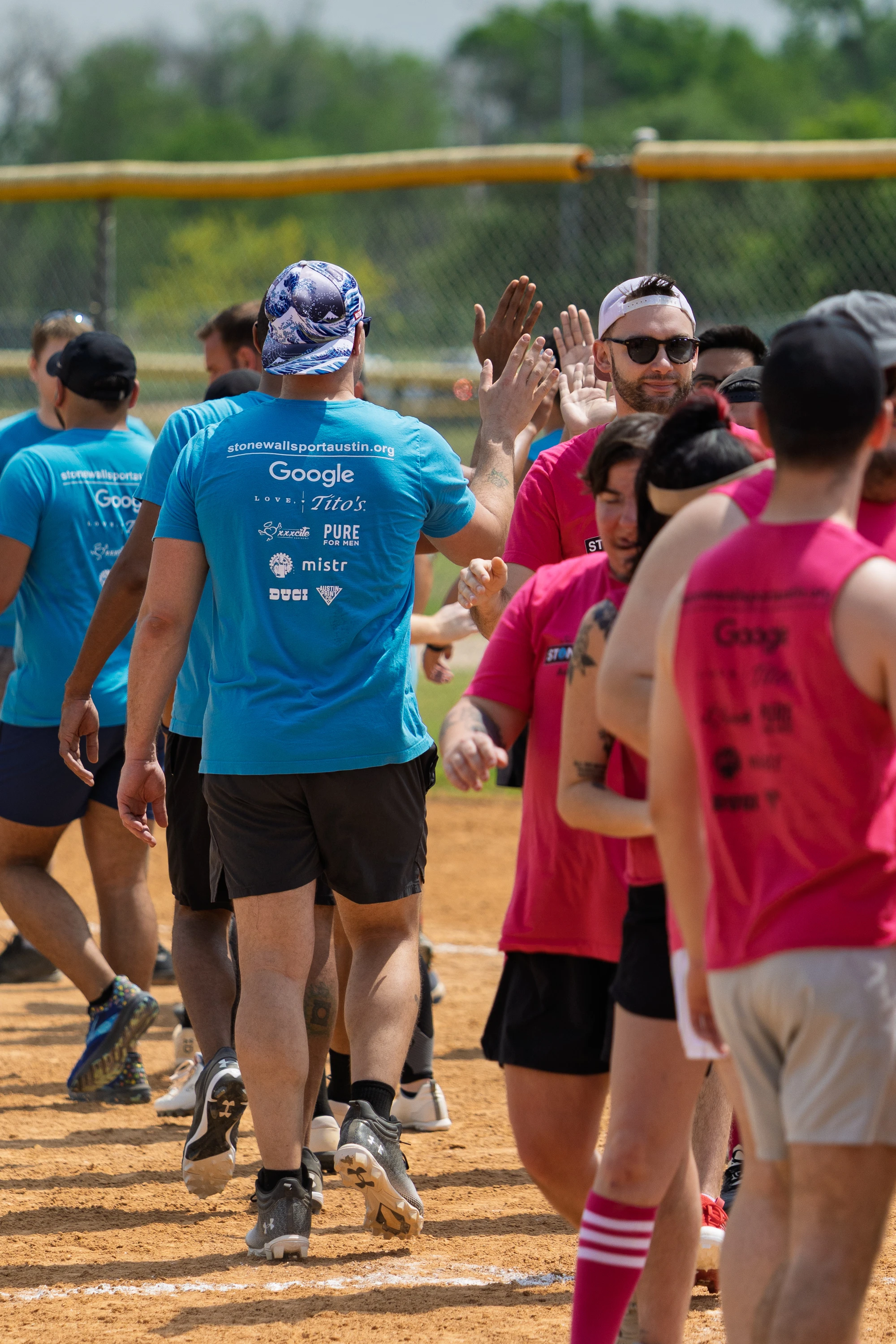 Players wearing blue and pink team shirts are lined up on a kickball field, giving each other high fives and handshakes after a game. The scene takes place outdoors on a sunny day, with a dirt field and green trees in the background.