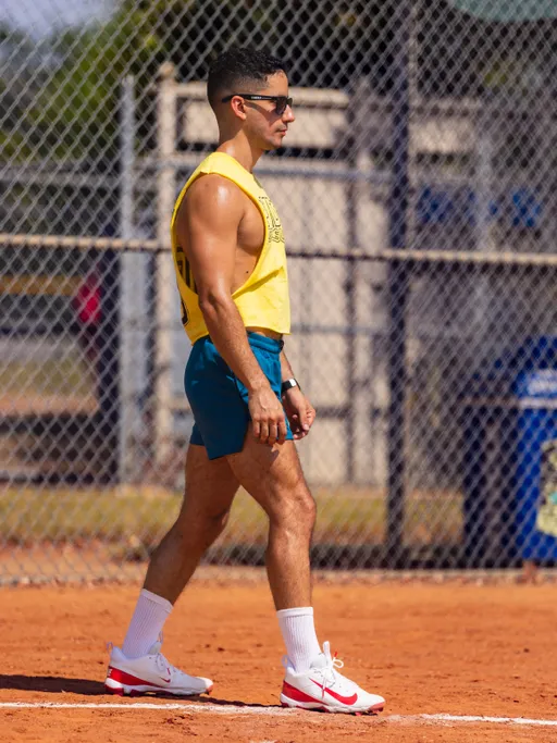 A kickball player walks along the dirt baseline on a sunny field, wearing a yellow team tank with text, teal shorts, white crew socks, and white sneakers with red accents, plus sunglasses and a watch; a chain-link fence and dugout area are in the background.