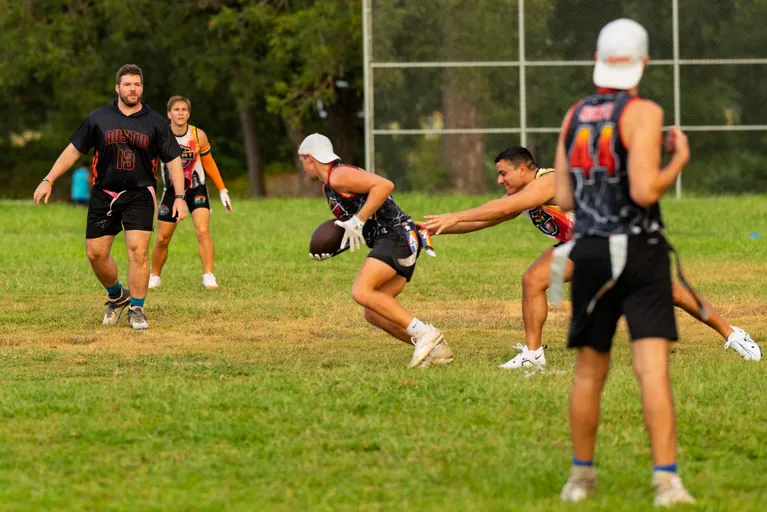 During practice for Gay Bowl 2025, ATX Heat (Orange) players run a flag-football drill on a grassy field: a ball carrier in a white cap and black jersey with orange accents cuts past a defender reaching for the flag belt, while teammates in numbered jerseys and cleats close in; trees and a chain-link fence frame the background.