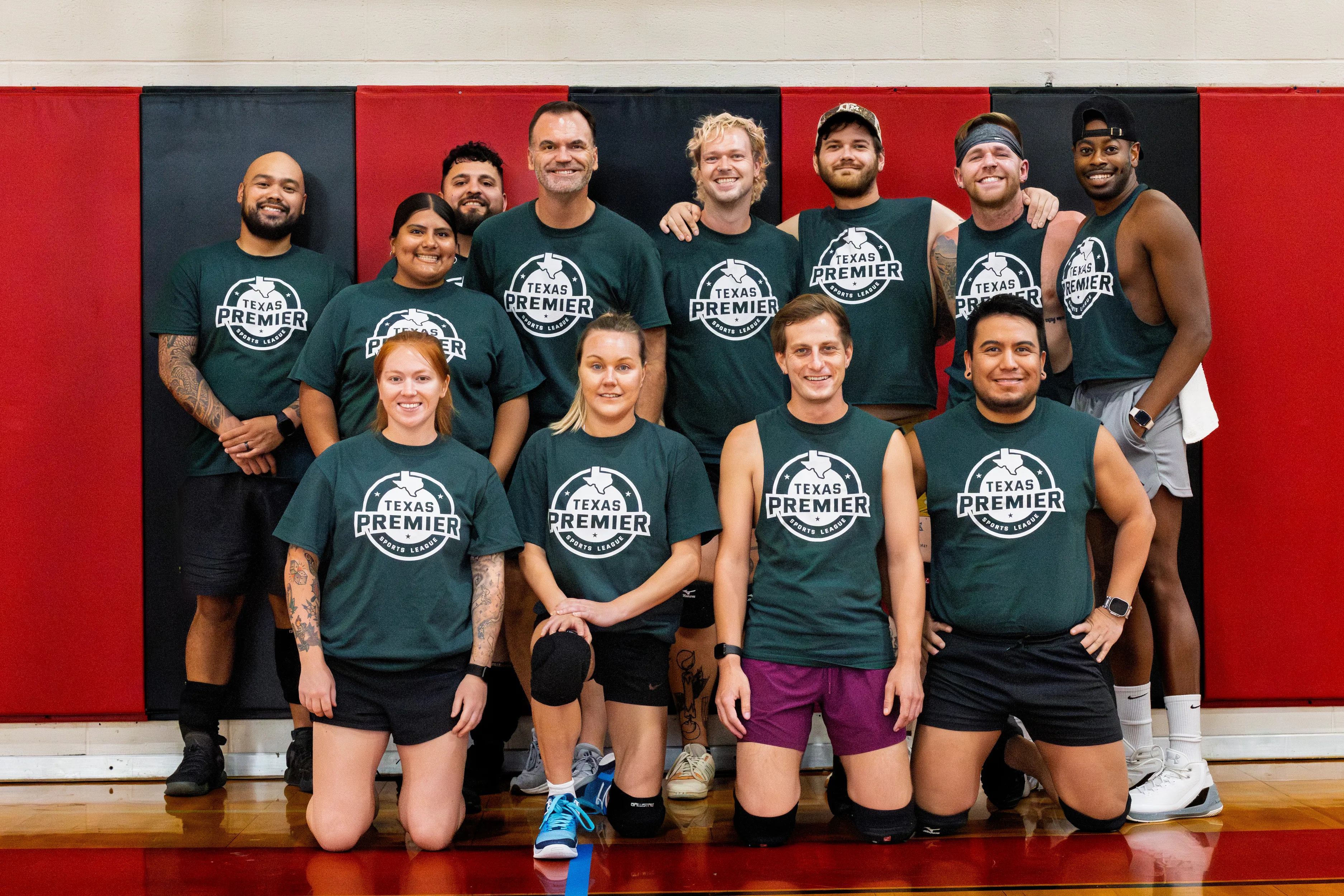 TPSL Dodgeball Summer 2025 team photo: a group of players in matching dark green “Texas Premier” shirts poses on an indoor dodgeball gym floor, some kneeling in front and others standing with arms around teammates. They wear athletic shorts and sneakers, with a few knee pads, headbands, and a cap visible, set against red-and-black wall padding.