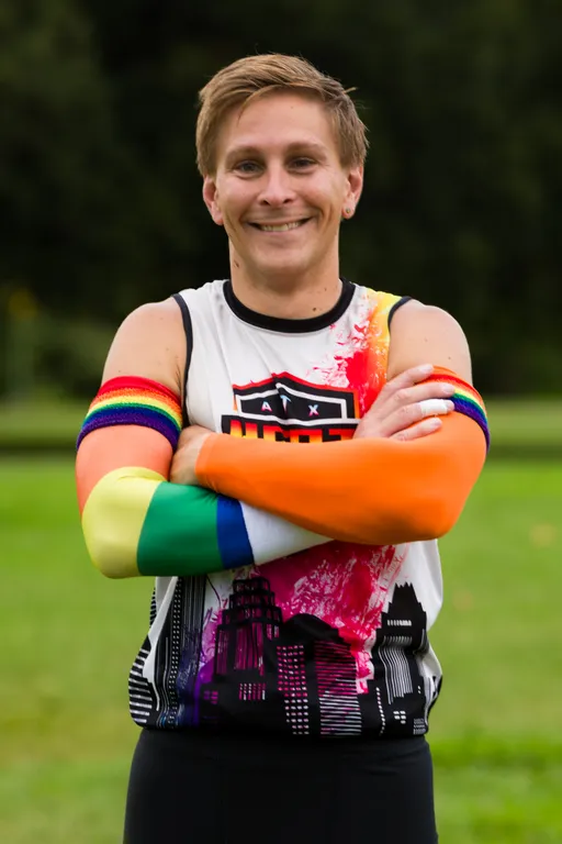 An ATX Heat (Orange) player stands on a grassy practice field with arms crossed, wearing a white sleeveless team jersey featuring the ATX Heat logo and a city skyline graphic, plus rainbow-striped and orange compression arm sleeves, with trees in the background during Gay Bowl 2025 practice.