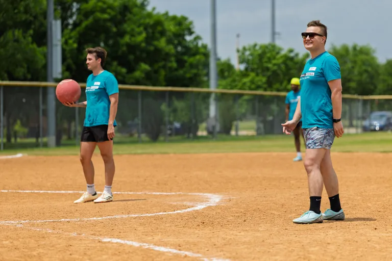 Two kickball players wearing matching teal shirts and shorts stand on a dirt field; one player is holding a red kickball while the other smiles and faces forward, with trees and a fence visible in the background.