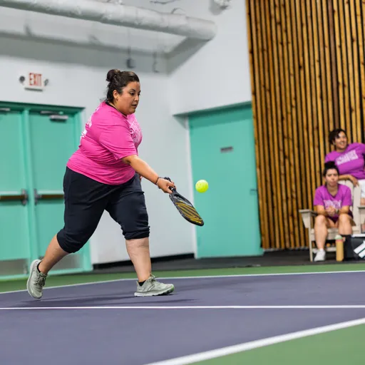 A player wearing a bright pink shirt, black pants, and gray shoes is mid-action on an indoor pickleball court, swinging a paddle to hit a ball; in the background, other players or supporters watch from sidelines near teal doors and wood-paneled walls.