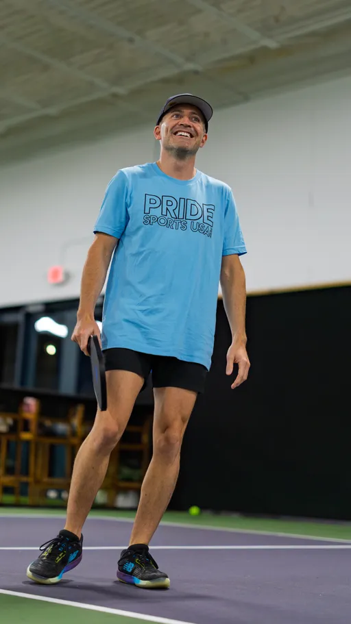 A pickleball player on an indoor court is smiling and holding a paddle, wearing a light blue "Pride Sports USA" shirt, black shorts, a cap, and athletic shoes. The player appears relaxed between points, with part of the court and some equipment visible in the background.