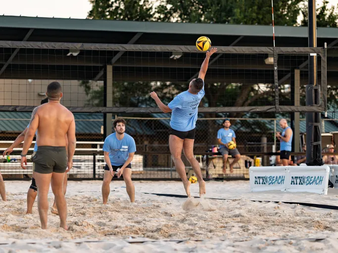 A volleyball player in a blue shirt is jumping to spike the ball over the net on a sand court, while other players watch and prepare for the next move. The setting is an outdoor beach volleyball facility with teammates and spectators in the background.