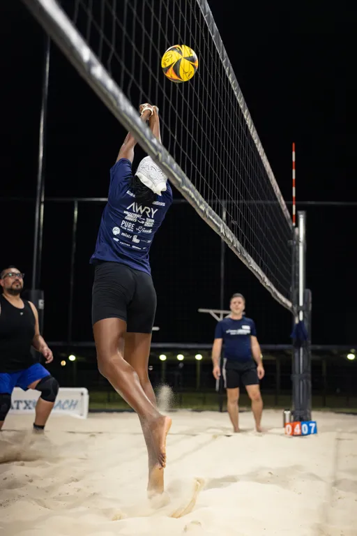 A player in a navy team shirt and black shorts leaps at the net, arms fully extended to block or tip a yellow-and-black volleyball as sand sprays beneath their feet under the night lights. Other players look on from the sand court, with a scoreboard by the post showing 04–07.
