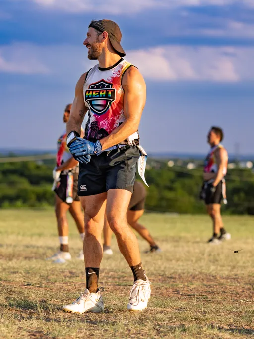 ATX Heat (Black) players run a flag football practice for Gay Bowl 2025 on an open grassy field; a foreground player in a white-and-red HEAT jersey, black shorts, backward cap, blue gloves, cleats, and a flag belt pauses between plays while teammates in matching gear reset in the background under a bright, late-day sky.