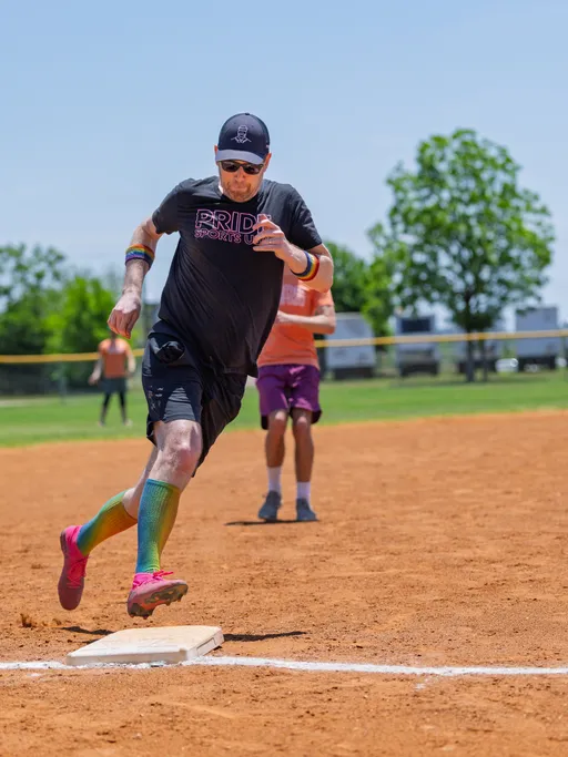A kickball player wearing a "Pride Sports" shirt, rainbow knee-high socks, and a cap is running and about to tag a base on a sunny, outdoor kickball field, while other players are visible in the background.