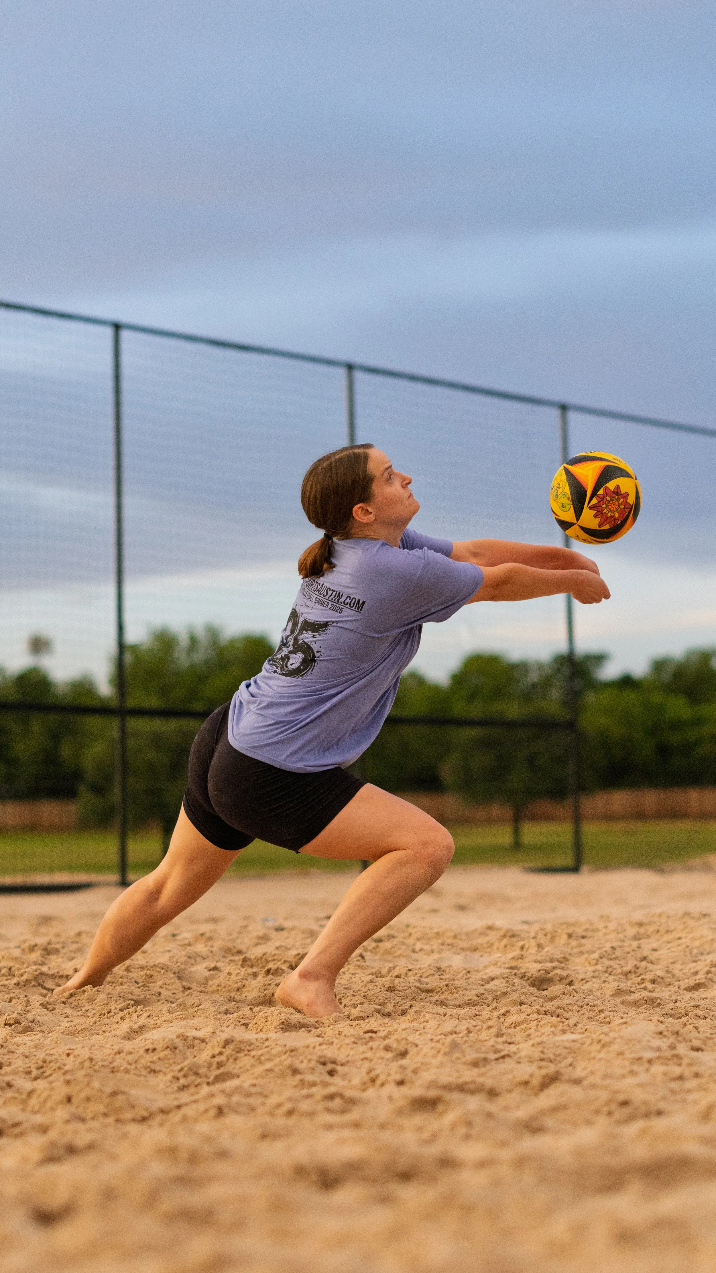 A player in a light purple team shirt with a printed number and black shorts lunges low in the sand, arms extended to bump a yellow-and-red volleyball. The action takes place on an outdoor sand court bordered by fencing and trees under an overcast sky.