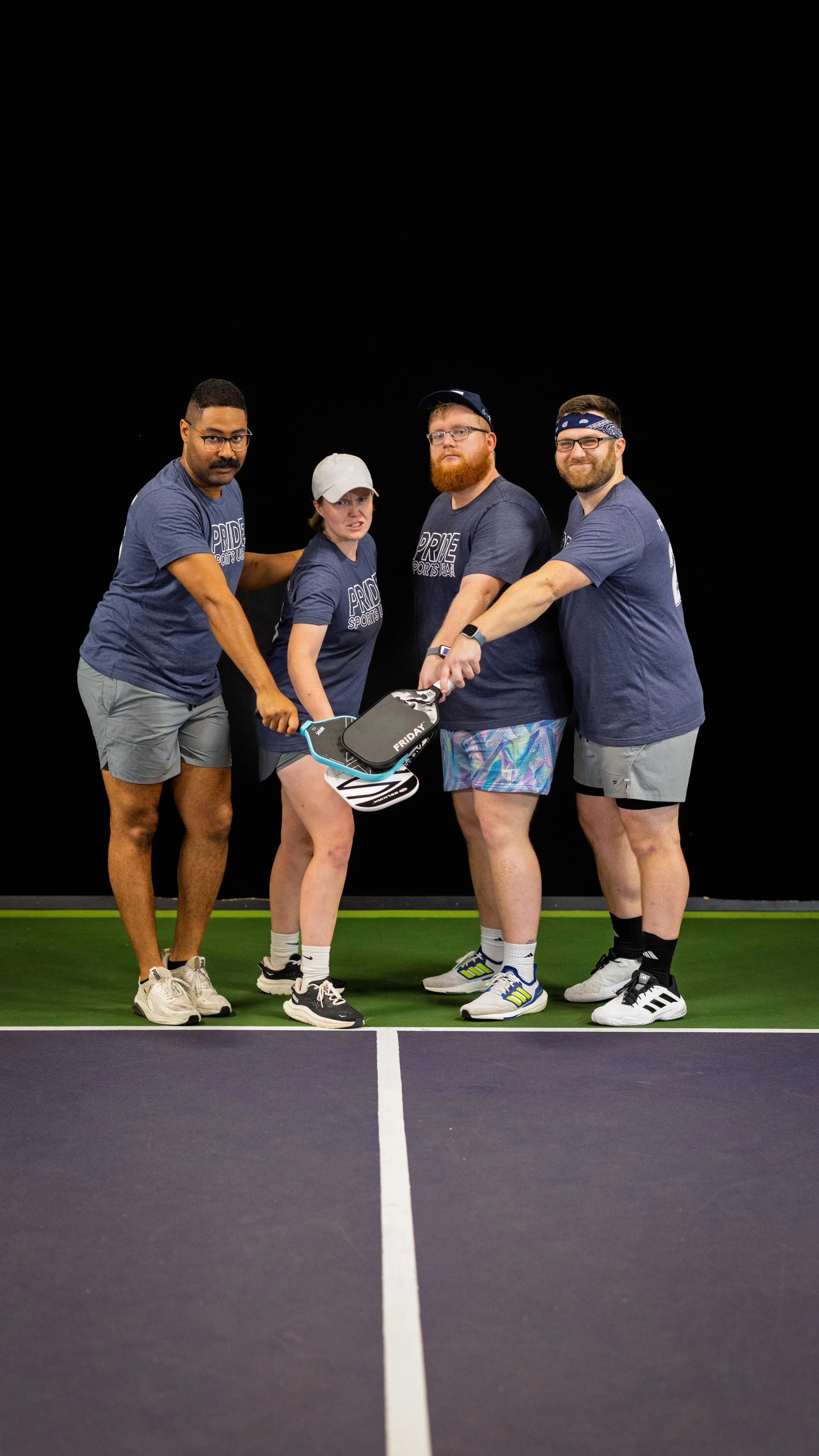 Four pickleball players stand together on an indoor court, each wearing matching athletic shirts and shorts, holding their paddles in the center with their hands stacked in a team gesture. The group appears ready and unified, with the paddle court lines visible in the foreground.