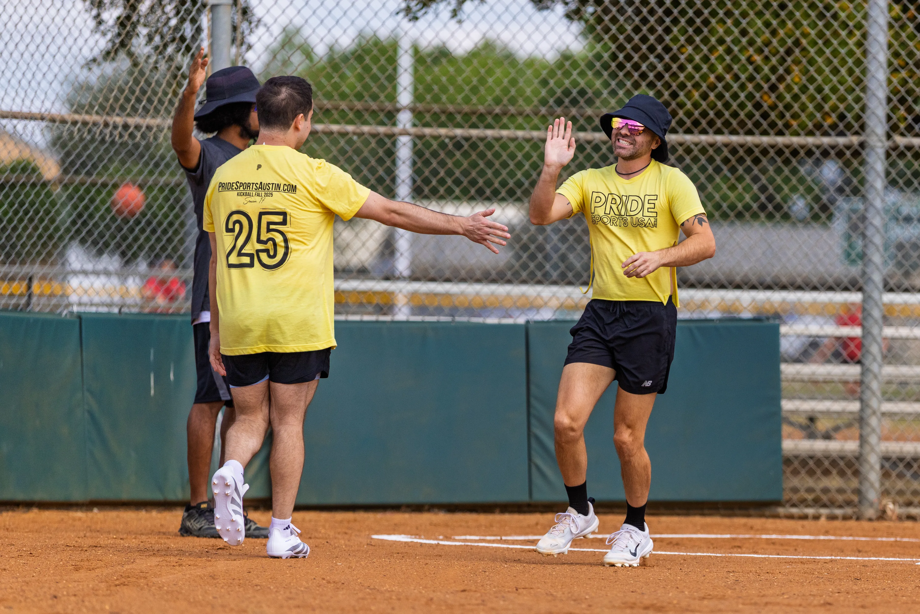 On a kickball field, three Pitch Please players in matching yellow PRIDE shirts celebrate at home plate with a high-five, one jersey showing the number 25 and “pridesportsaustin.com.” A chain-link backstop with green padding and bleachers frames the scene, a bright orange kickball is blurred in the background, and some players wear bucket hats, sunglasses, and athletic shoes.