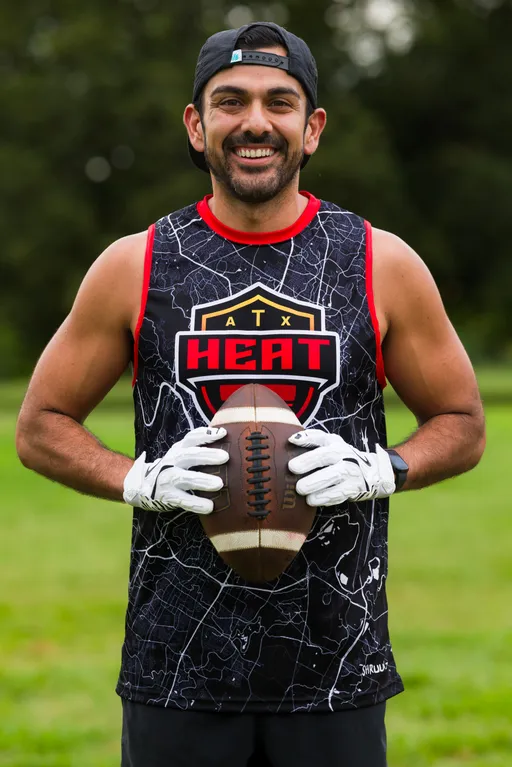 An ATX Heat player stands on a grassy field during practice for Gay Bowl 2025, smiling and holding a football at chest height while wearing a black map-pattern sleeveless jersey with red trim and an “ATX HEAT” crest, white receiver gloves, a backward cap, and a wristwatch; trees blur in the background.