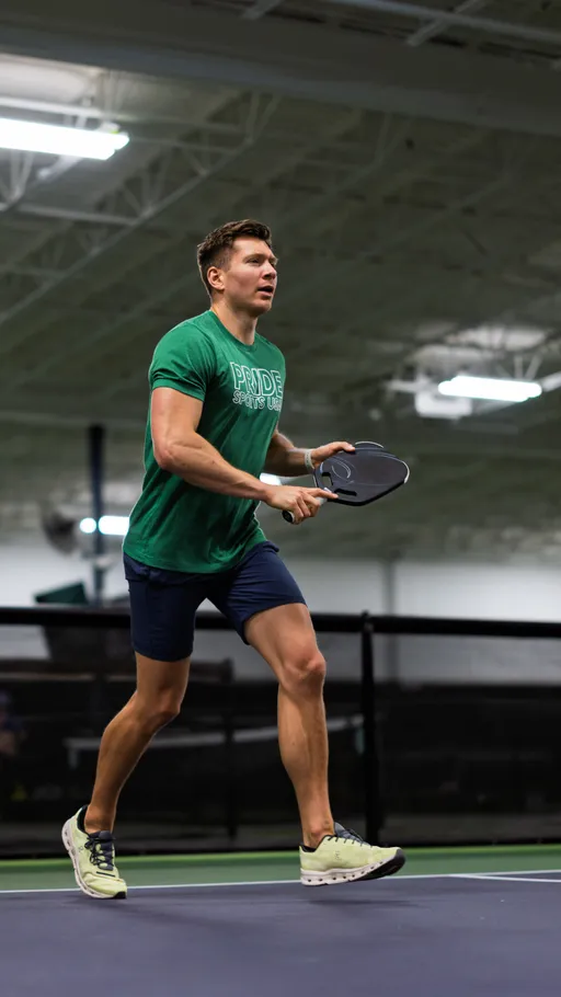 A pickleball player wearing a green "Pride Sports League" t-shirt and blue shorts prepares to hit a ball on an indoor court, holding a paddle while in motion.