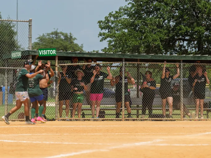 Two kickball players wearing athletic clothing and sneakers celebrate near home plate while members of their team cheer enthusiastically from behind the chain-link fence in the dugout labeled "Visitor"; the scene takes place on a sunny outdoor kickball field with green trees in the background.