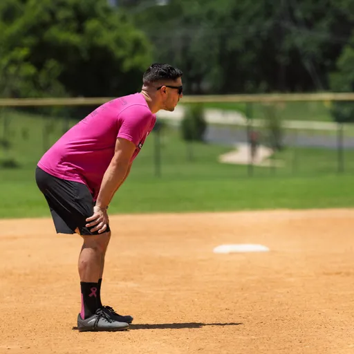 A kickball player wearing a bright pink shirt, black shorts, and matching pink socks is positioned on a dirt field, slightly bent forward in a ready stance with hands on knees, appearing focused during a game.