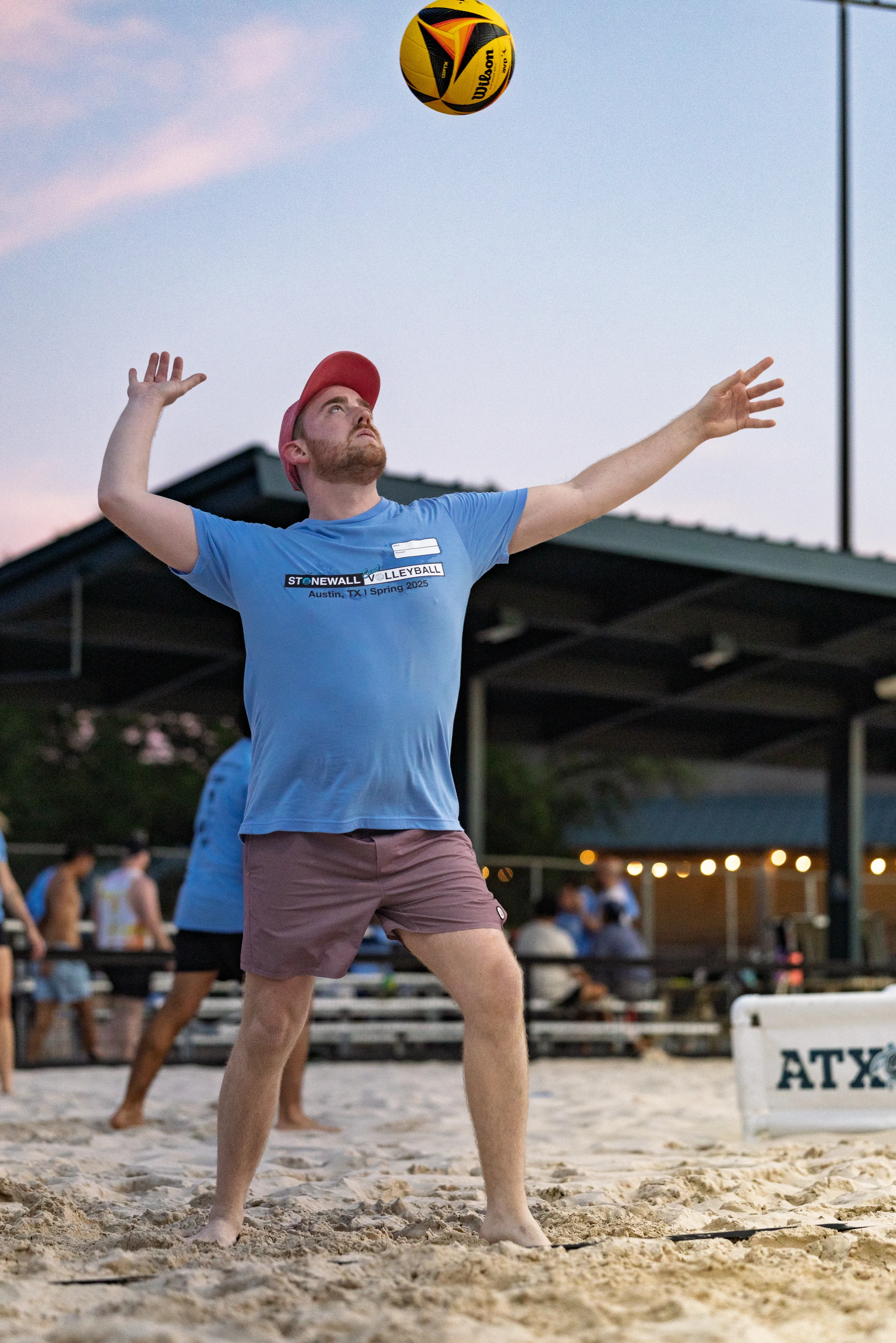 A volleyball player wearing a red cap, blue Stonewall Volleyball t-shirt, and shorts is preparing to serve the ball on a sandy outdoor court, with a pavilion and other participants in the background.