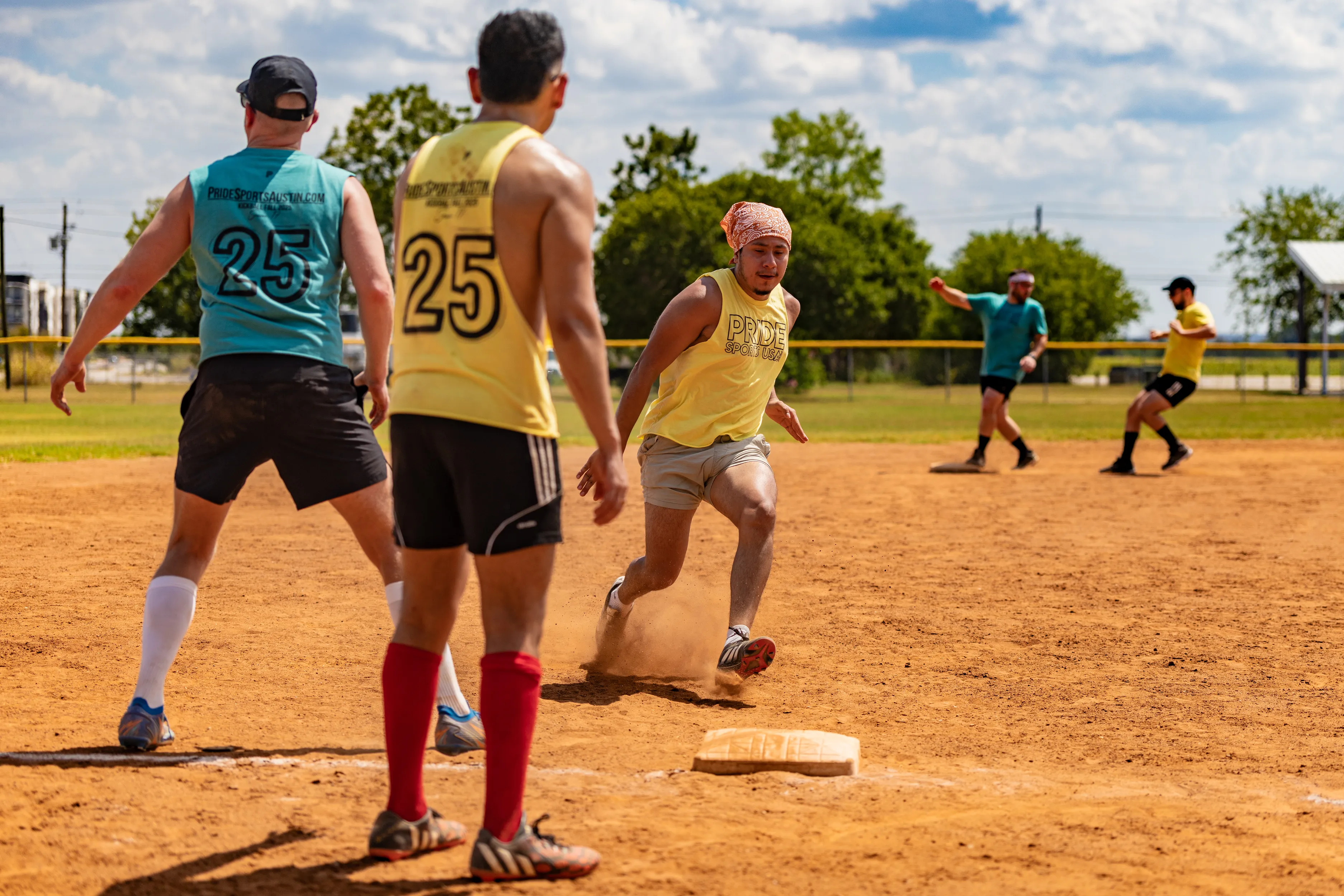 On a sunny kickball field, a player in a yellow jersey sprints through a base as dust kicks up, while teal- and yellow-clad defenders move in; numbers, knee-high socks, and headbands stand out against a grassy outfield and chain-link fence.