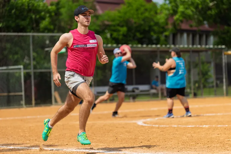 A player wearing a red "Stonewall Kickball" tank top, light shorts, and teal athletic shoes is running on a kickball field while another player in a blue shirt prepares to throw a red kickball; additional players and a chain-link fence are visible in the background.