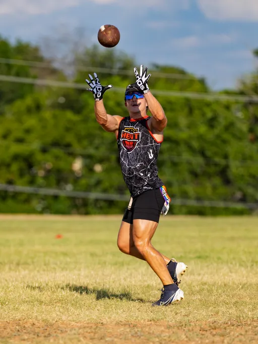 An ATX Heat (Black) player on a grassy practice field reaches up to catch a flag football pass, hands spread in patterned receiver gloves. The athlete wears a black HEAT jersey with orange trim, black shorts, cleats, blue sport sunglasses, and a flag belt, with trees and scattered cones in the background under bright sun.