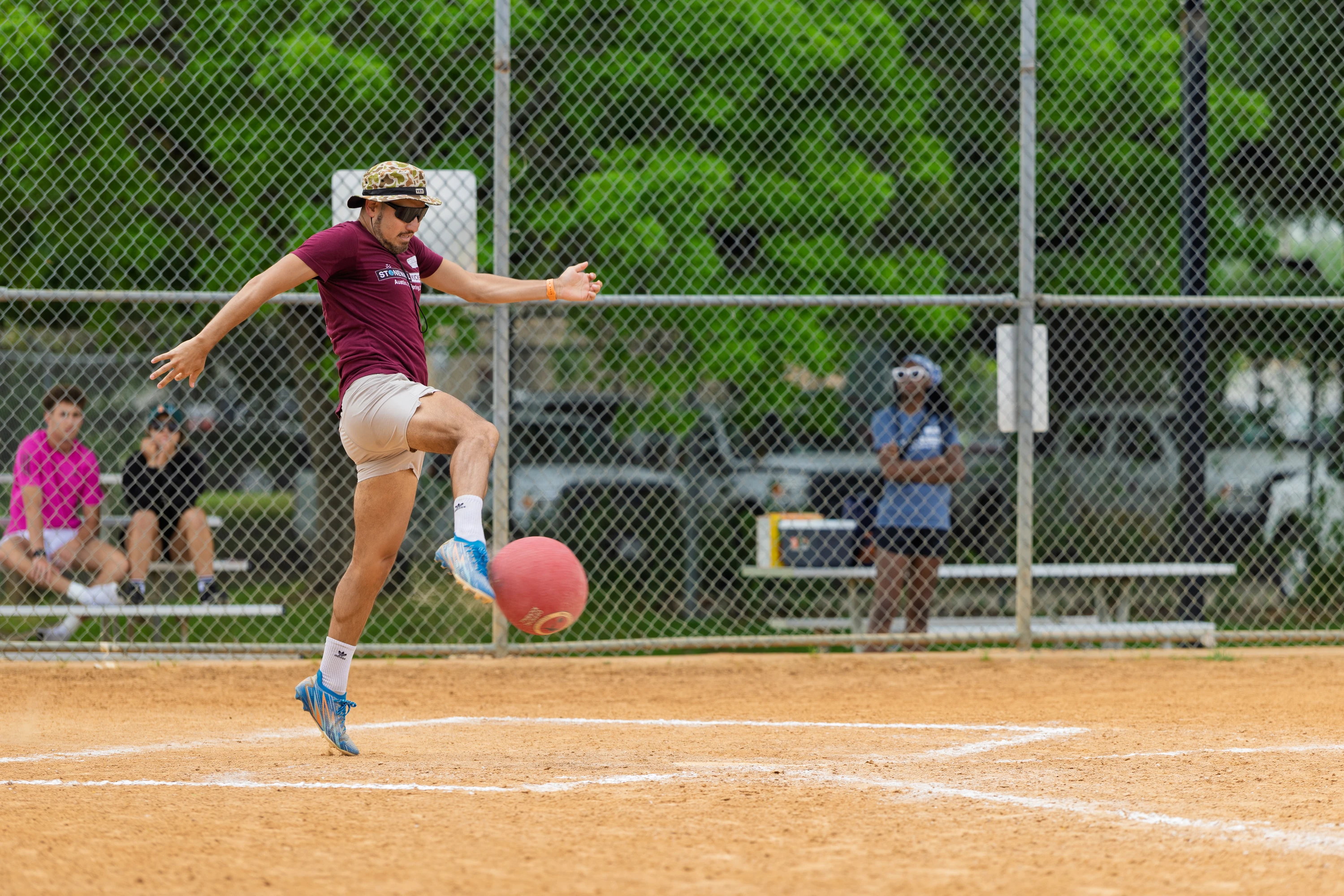 A kickball player in a maroon shirt, light shorts, and patterned hat is captured mid-kick on a sandy kickball field, while several people watch from behind a chain-link fence.