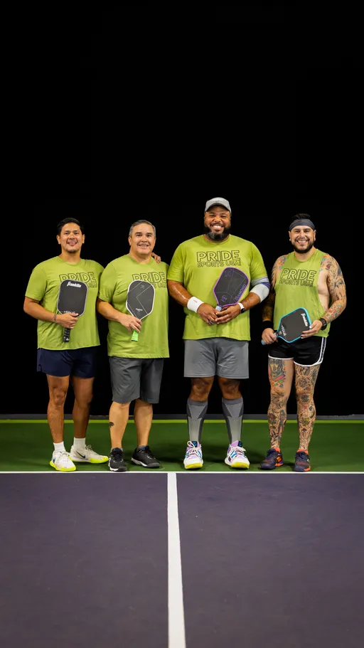Four pickleball players stand side by side on an indoor court, each holding a paddle and wearing matching green “Pride Sports USA” shirts; one player is notable for having arm and leg tattoos, a headband, and shorts.