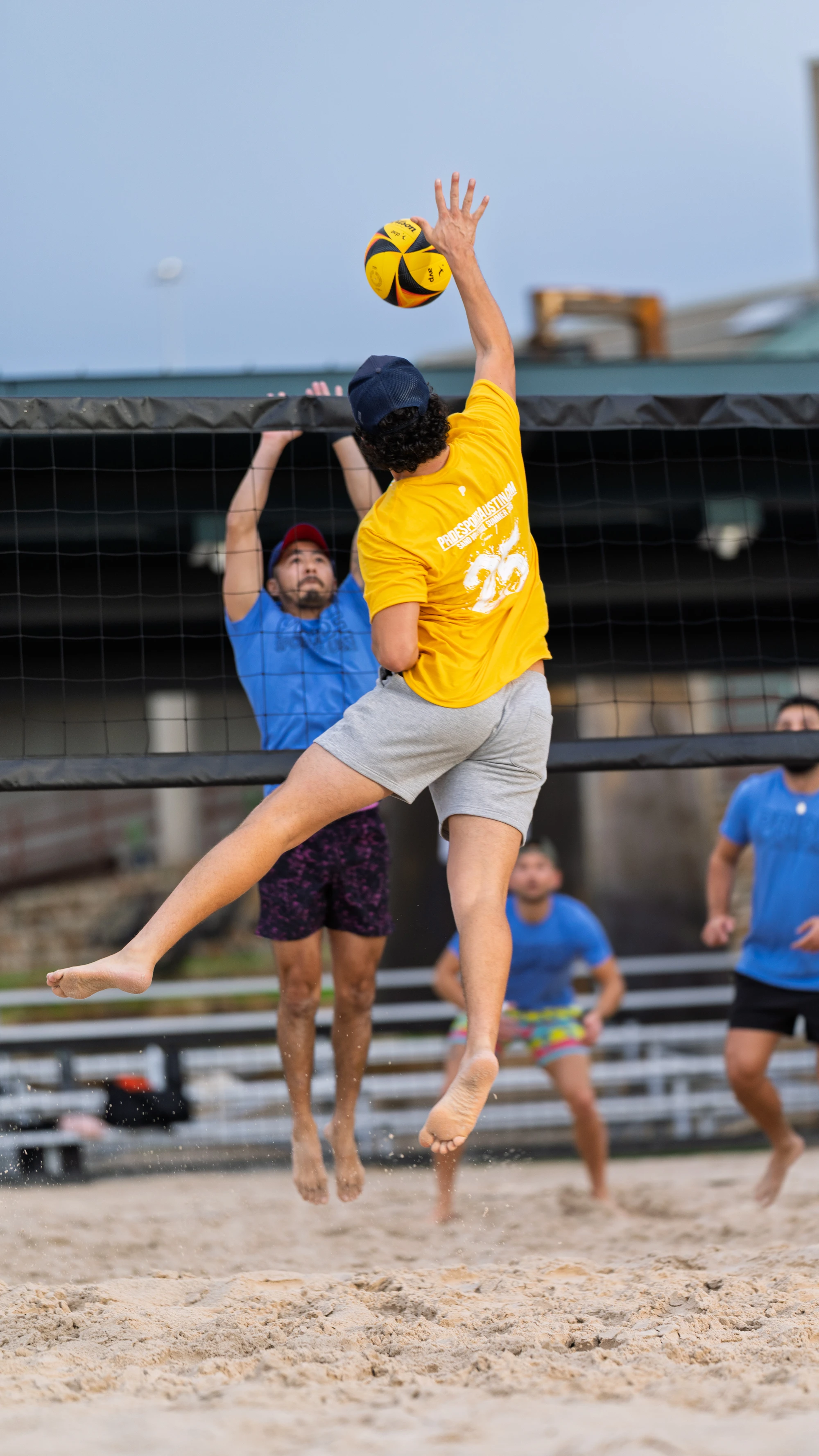 A volleyball player in a yellow shirt and cap leaps to spike the ball over the net, while an opposing player in a blue shirt and hat jumps to block; other teammates stand ready on the sandy outdoor court.