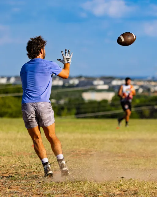 On a dry grassy field, an ATX Heat (Black) player in a blue shirt, gray camo shorts, gloves, and cleats reaches forward to catch a spiraling football, kicking up dust. Another player with a flag belt runs in the background against a distant cityscape, suggesting a passing drill during Gay Bowl 2025 practice.