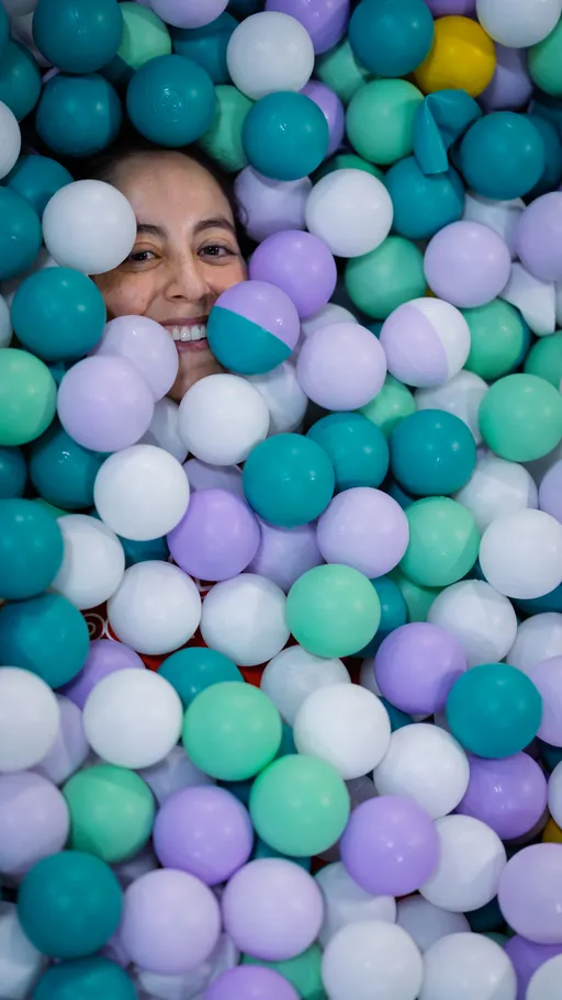 A person is partially submerged and smiling in a pit filled with colorful plastic balls in shades of teal, lavender, white, and a few yellow, suggesting a playful and joyful moment during the Pride Pickleball Summer 2025 event.