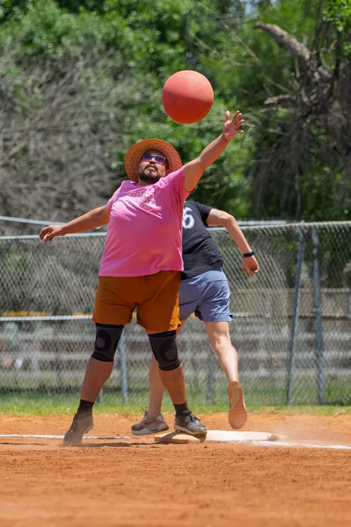 A player wearing a wide-brimmed hat, sunglasses, a pink shirt, and brown shorts jumps to reach for a kickball in mid-air on a sandy kickball field, with another player running behind them.