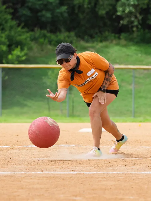 A kickball player wearing an orange team shirt, black cap, and sunglasses is in the act of pitching the ball on a sandy kickball field, with green grass and a fence in the background.