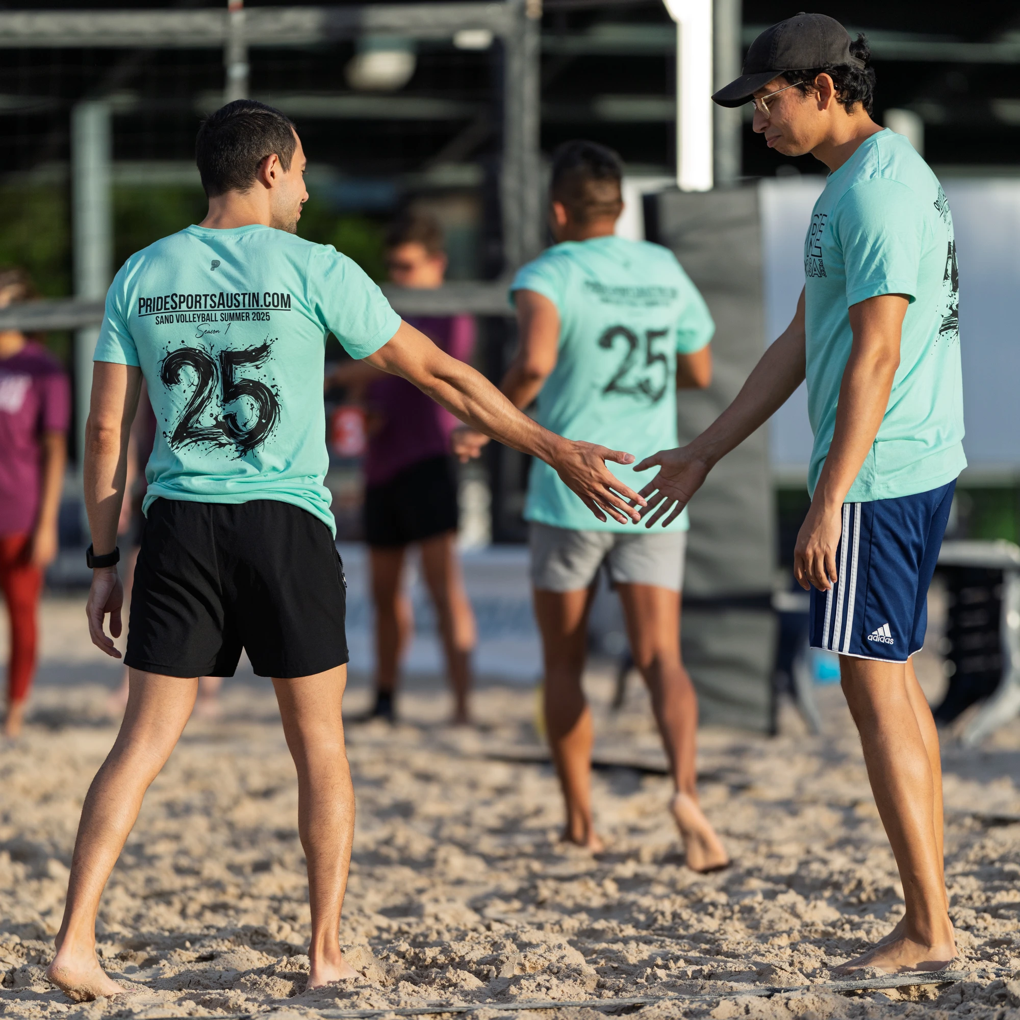 Players wearing matching turquoise shirts and athletic shorts are on a sand volleyball court, giving each other high-fives after a play. The scene is set outdoors with a volleyball net in the background, and the group appears to be celebrating or encouraging one another.