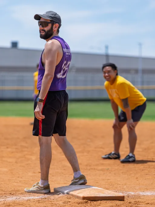 A player wearing a purple tank top, black shorts, and a cap stands on a base on a kickball field, while another player in a yellow shirt and black shorts stands in the background, appearing to watch the action and smiling.