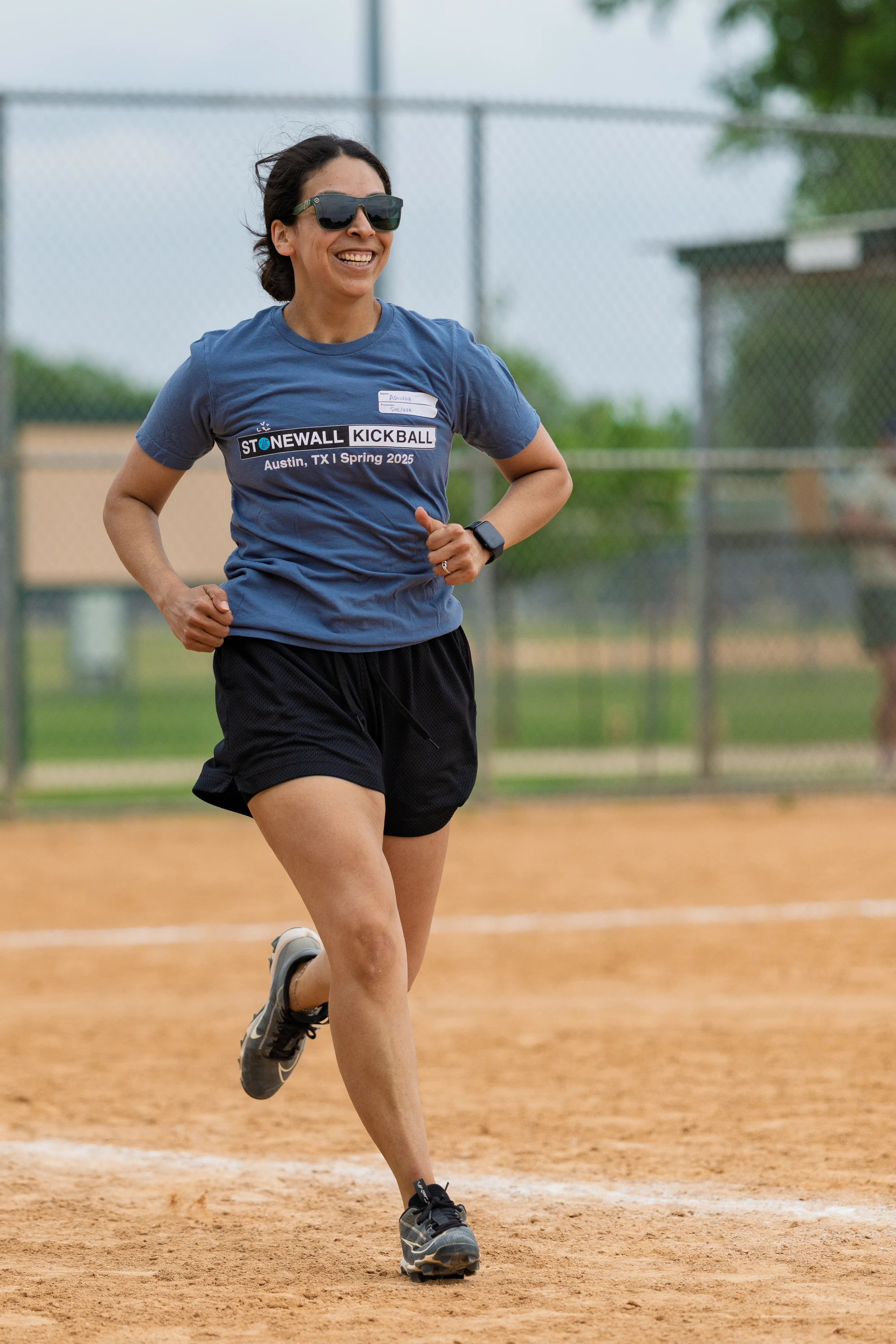 A player wearing sunglasses, a "Stonewall Kickball Austin, TX | Spring 2025" t-shirt, black shorts, and sports shoes is running on a kickball field, smiling during the game.