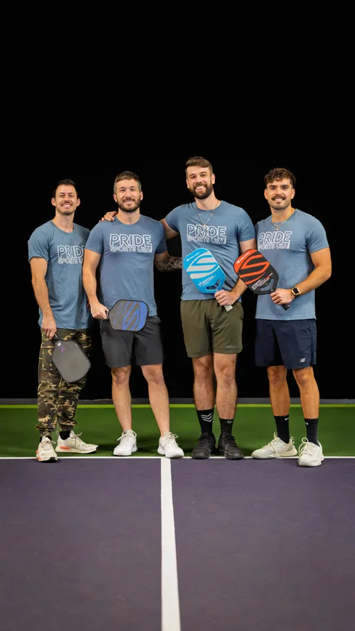 Four pickleball players stand together on an indoor court, each holding a pickleball paddle and wearing matching “PRIDE SPORTS USA” t-shirts; the group appears relaxed and smiling, dressed in athletic shorts and sneakers.
