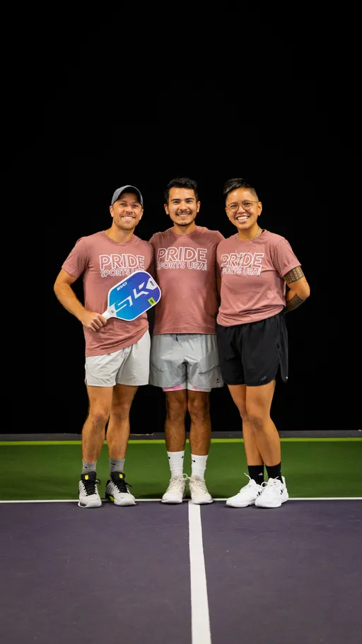 Three pickleball players pose together and smile on a court, each wearing matching "PRIDE SPORTS USA" t-shirts and athletic shorts, with one person holding a pickleball paddle.