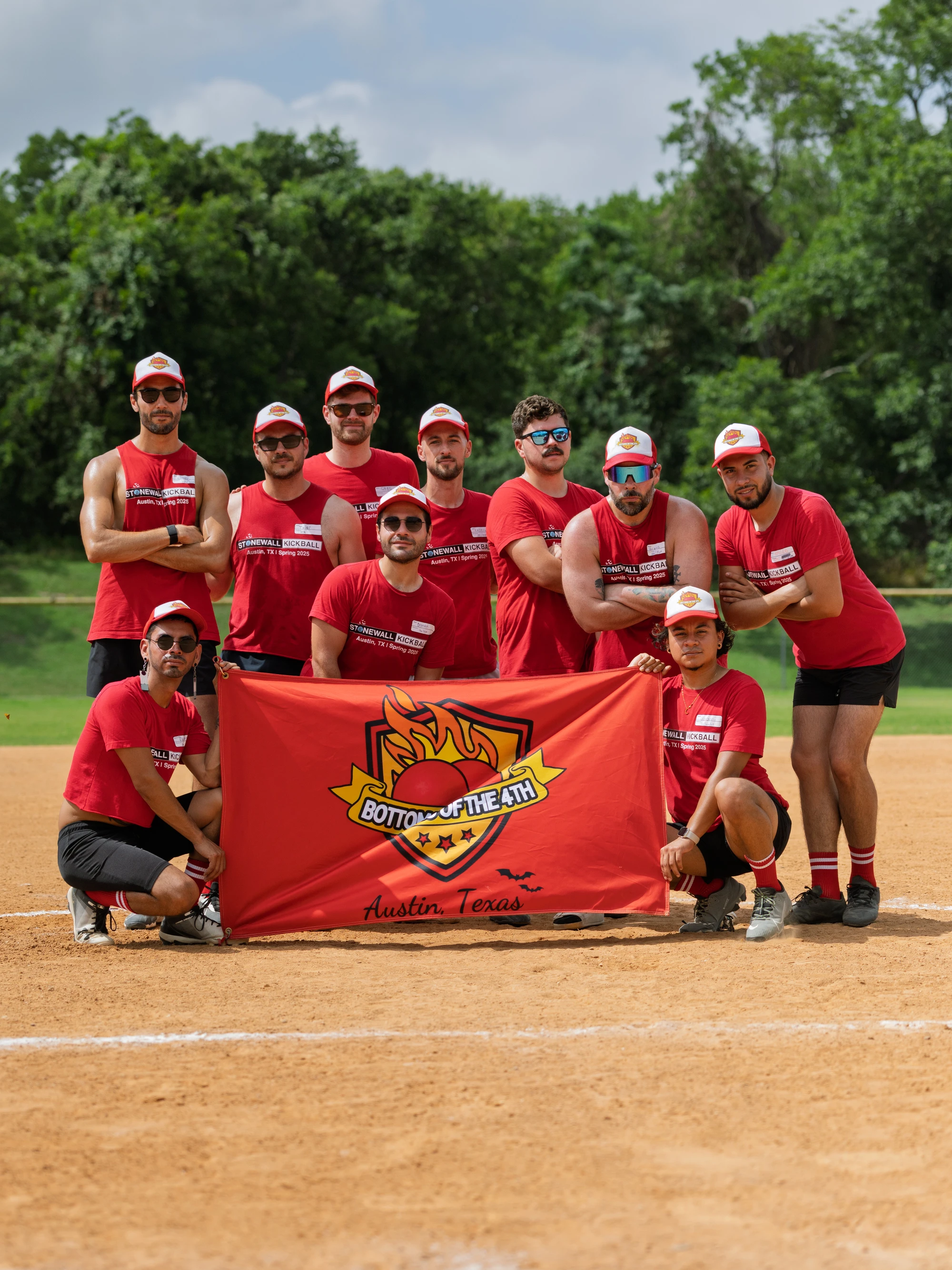 A group of kickball players wearing matching red shirts, black shorts, and red-and-white hats pose together on a dirt kickball field, holding a bright red team banner that reads "Bottom of the 4th, Austin, Texas" with a flame graphic. Lush green trees are visible in the background.