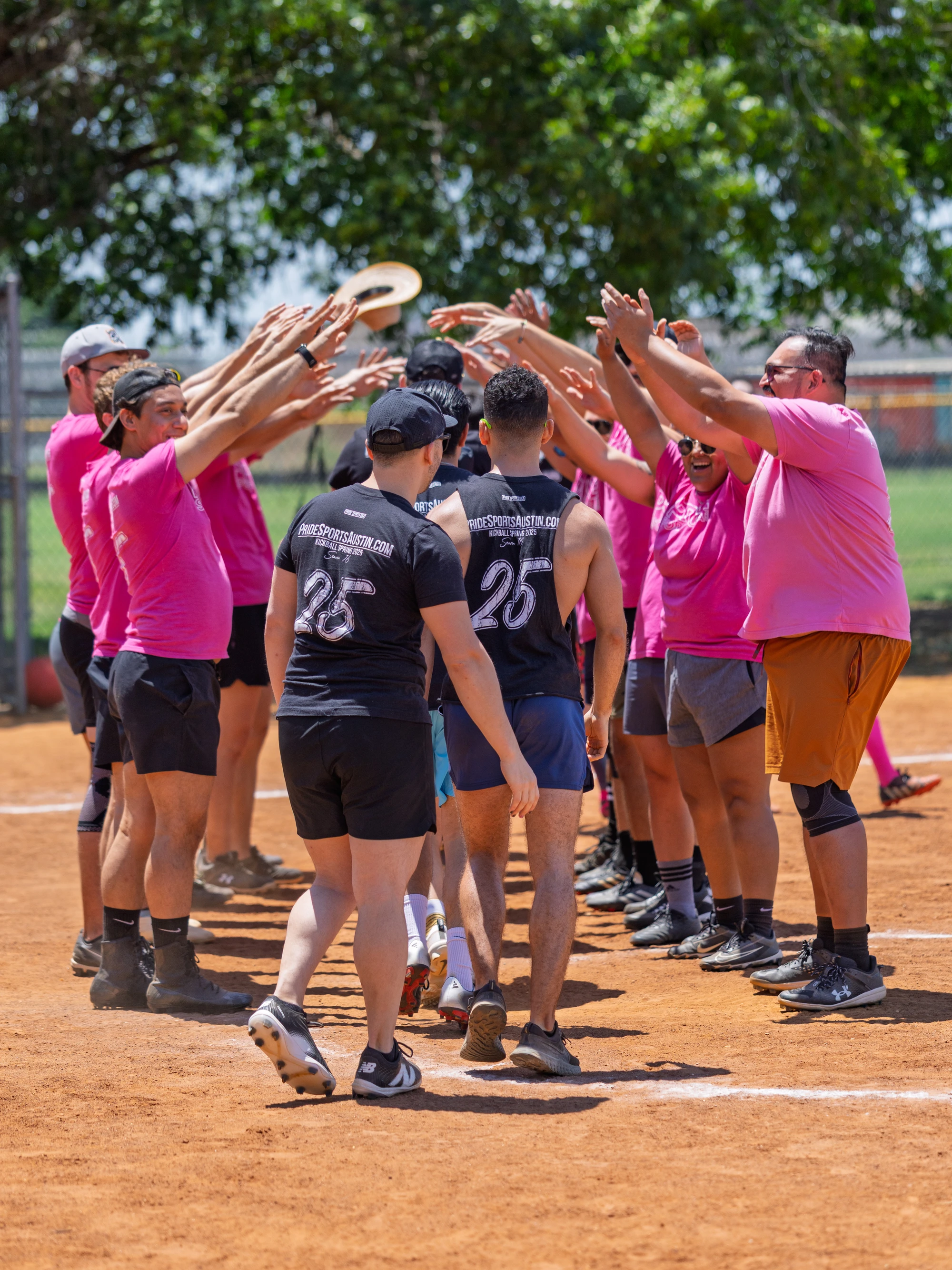 Players wearing bright pink and dark uniforms form two lines on a kickball field, creating an arch with their raised arms while others walk underneath, celebrating together in a festive outdoor setting.