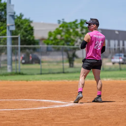 A kickball player wearing a bright pink jersey, black shorts, and a visor stands on the field, appearing to watch the game’s action with hands on hips. The player is on a dirt kickball field with grass and a chain-link fence in the background.