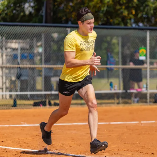 A player wearing a yellow “PRIDE SPORTS” shirt, black shorts, cleats, and a headband sprints on the red-dirt kickball field, kicking up dust as they cross a base line. The scene is a sunny park with a chain-link fence and blurred teammates and gear in the background.