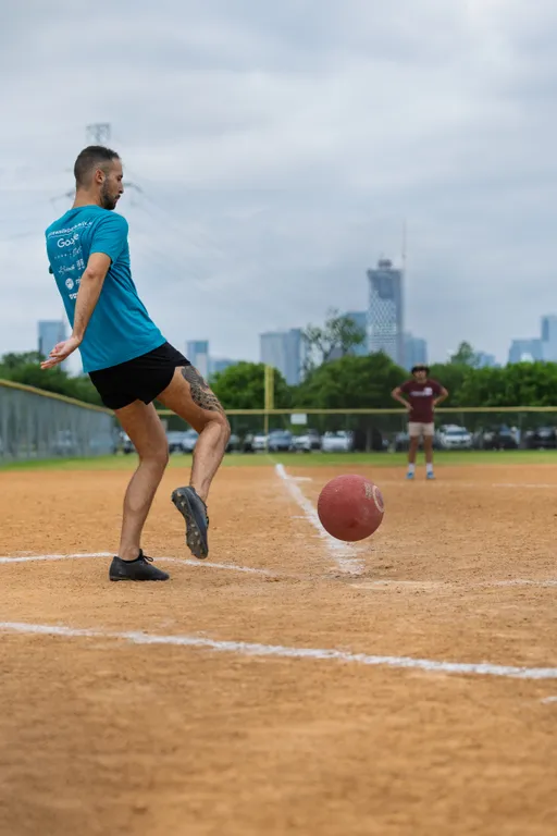 A player in a teal shirt and black shorts is kicking a red kickball on a dirt kickball field, with another player standing in the outfield and city buildings visible in the background.