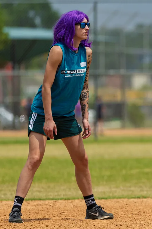 A kickball player with a bright purple wig, sunglasses, and a sleeveless Stonewall Kickball team shirt stands ready on a dirt section of the field, focused on the game. The player is wearing athletic shorts, socks, and cleats, with a tattoo visible on one arm, and the kickball field with fencing is visible in the background.