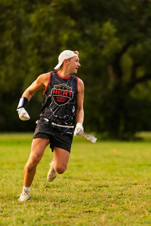 An ATX Heat (Orange) player sprints across a grassy field during Gay Bowl 2025 practice, head turned left as if tracking the play. The athlete wears a black HEAT jersey with orange trim, black shorts, a backward white cap, white gloves, cleats, a wrist playbook, and a flag belt with a patterned flag.