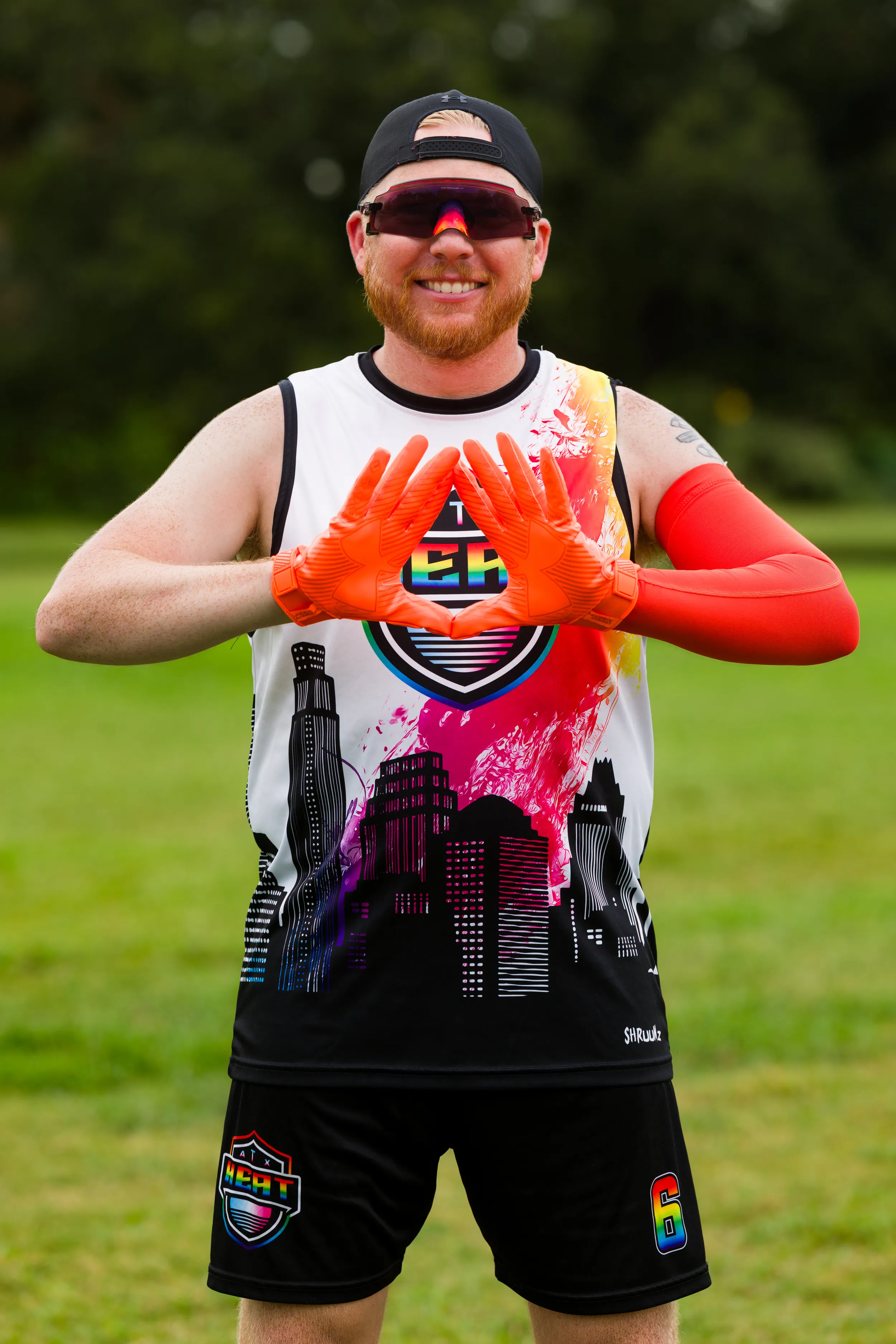 On a grassy practice field, an ATX Heat (Orange) player poses for Gay Bowl 2025 prep, wearing a black cap, mirrored sport shades, a sleeveless jersey with a colorful cityscape and team crest, black shorts with a crest and “6,” and bright orange receiver gloves and arm sleeve, holding gloved hands in a diamond shape.