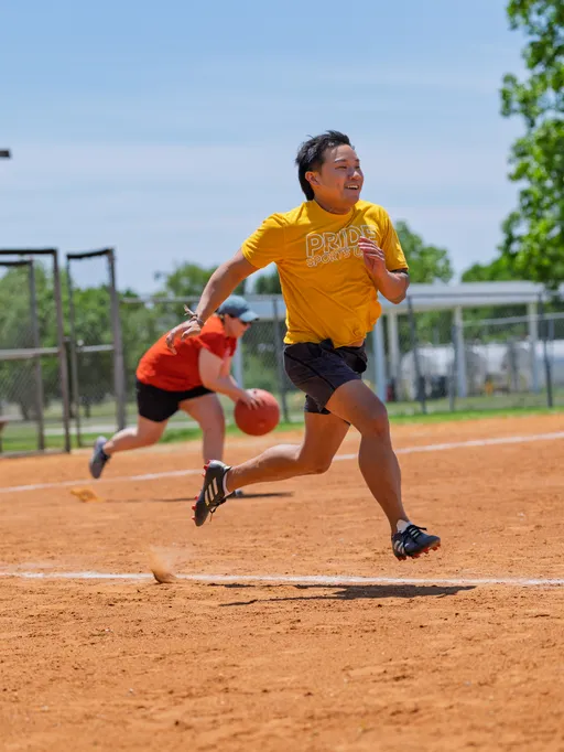 A player in a yellow "Pride Sports" shirt and black shorts is running across a kickball field, while another player in an orange shirt prepares to throw a kickball in the background. The scene is set outdoors on a sunny day with a fenced field and green trees visible.