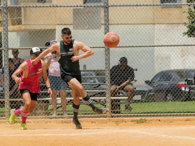 Two athletes are playing on a kickball field; one player wearing a green tank top and knee-high socks is kicking a red kickball, while another player in a pink jersey and cap runs beside them. A chain-link fence and spectators are visible in the background.