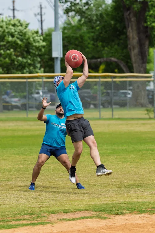 Two kickball players wearing blue shirts and athletic shorts are on a grassy field; one player is jumping high in the air to catch a red kickball above their head while the other player reaches up from behind, mid-action.