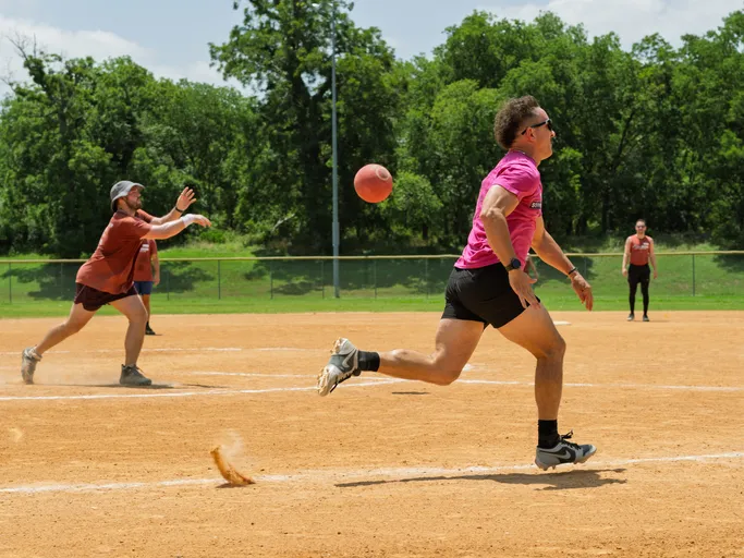 A player in a bright pink shirt and black shorts runs towards first base as a kickball rolls through the air, while another competitor in a red shirt and cap reaches out to catch it on a sunny outdoor kickball field.